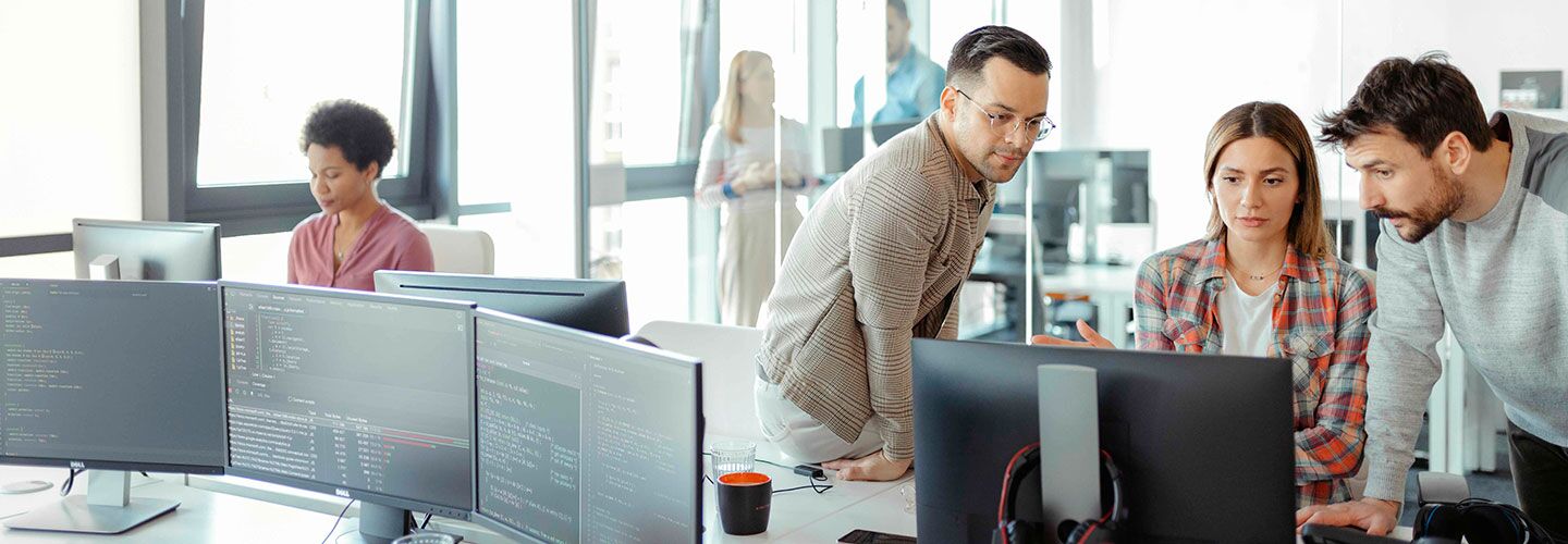 Team von drei Personen arbeitet gemeinsam vor einem Monitor, im Vordergrund mehrere Bildschirme mit Programmiercode. Eine weitere Person arbeitet konzentriert an einem Schreibtisch im Hintergrund, moderne Büroumgebung mit großen Fenstern und Glaswänden.